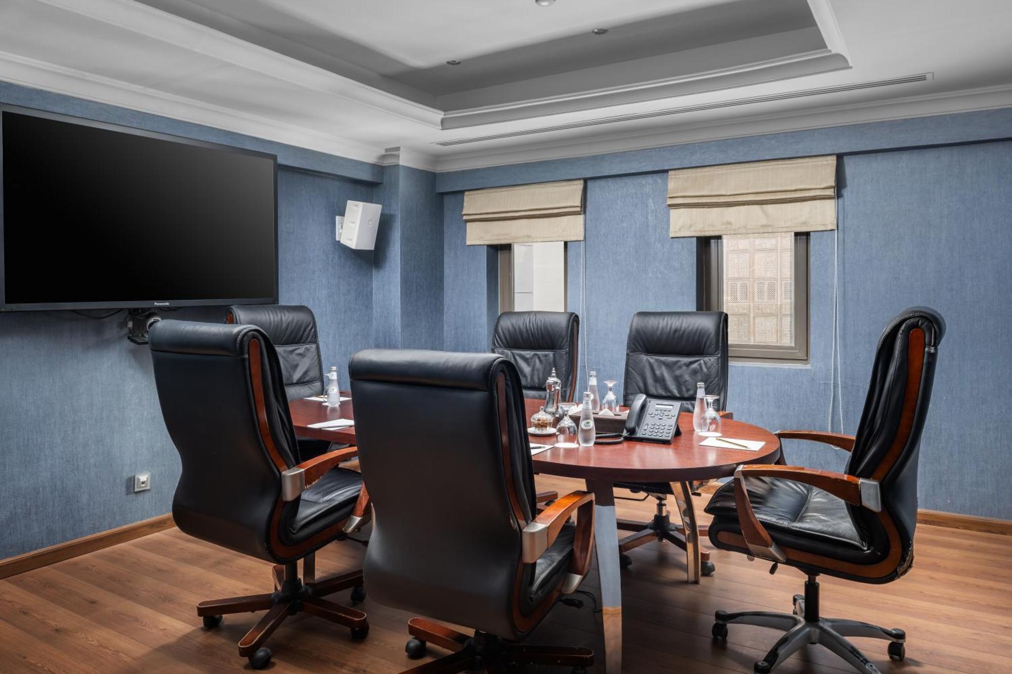 Al Aqeeq Madinah Hotel Medina Exteriör bild The photo shows a modern conference room. It features a round wooden table surrounded by several black leather swivel chairs. The walls have a blue color scheme, and there is a large flat-screen TV mounted on one wall. The room has two windows with b