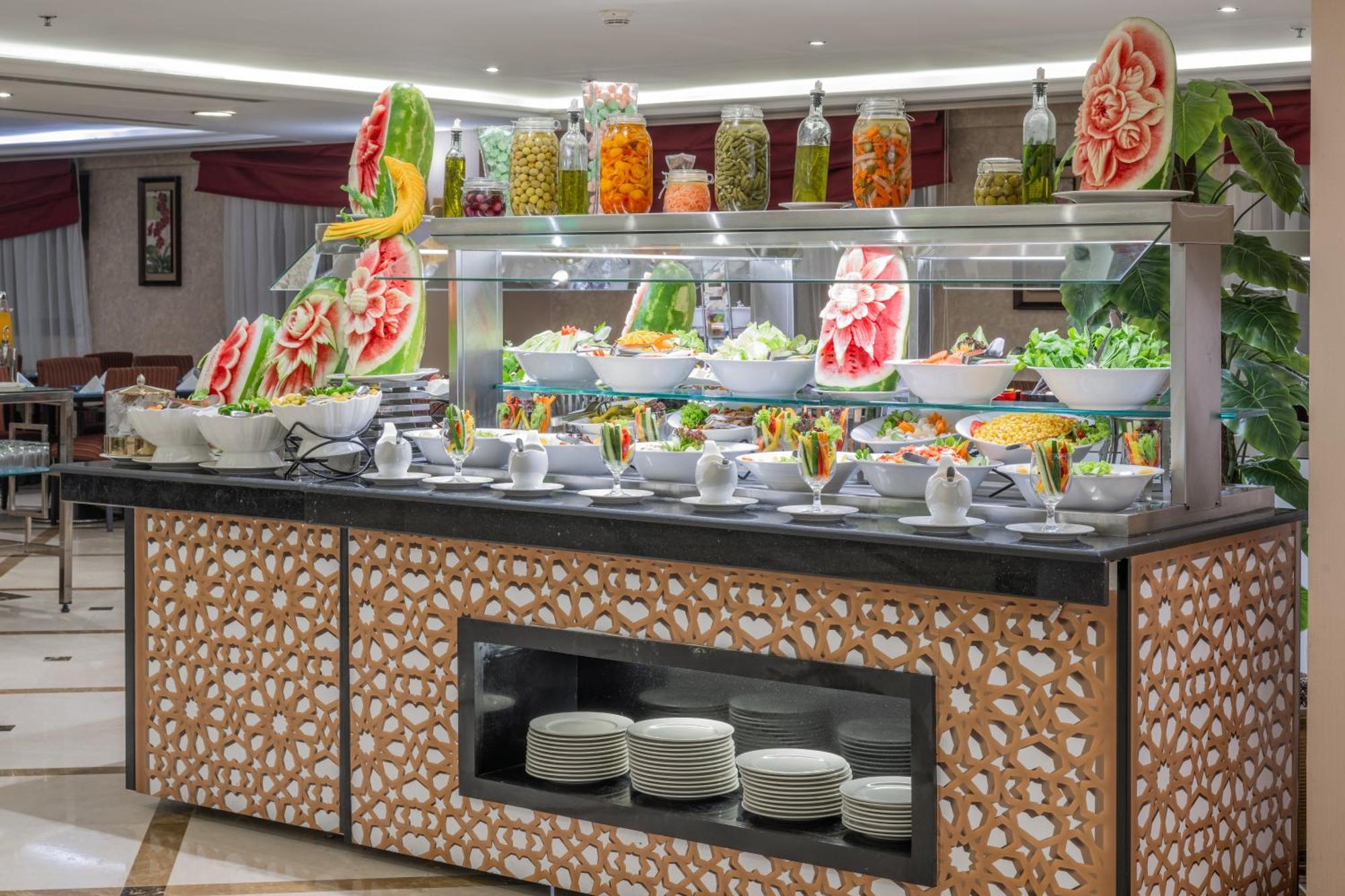Al Aqeeq Madinah Hotel Medina Exteriör bild The photo shows a buffet display that features an array of colorful food items. In the foreground, there are several large bowls filled with various salads and dishes. The presentation is visually appealing, with decorative watermelon carvings and an