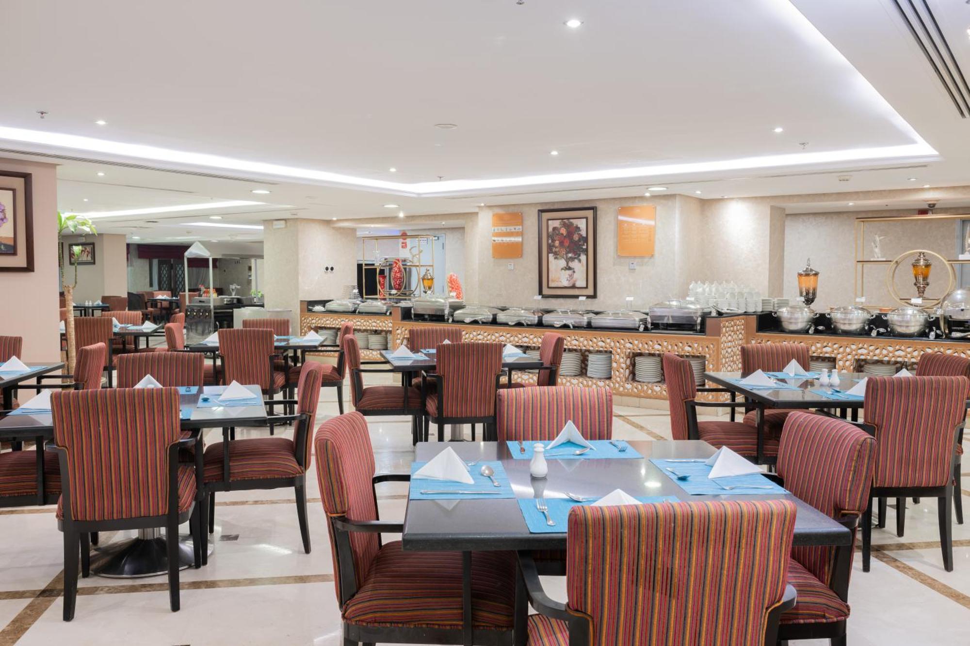 Al Aqeeq Madinah Hotel Medina Exteriör bild The photo shows the interior of a restaurant. It features multiple tables arranged neatly, each with patterned chairs and set with blue and white table settings, including folded napkins. The lighting is bright and modern, creating a welcoming atmosp
