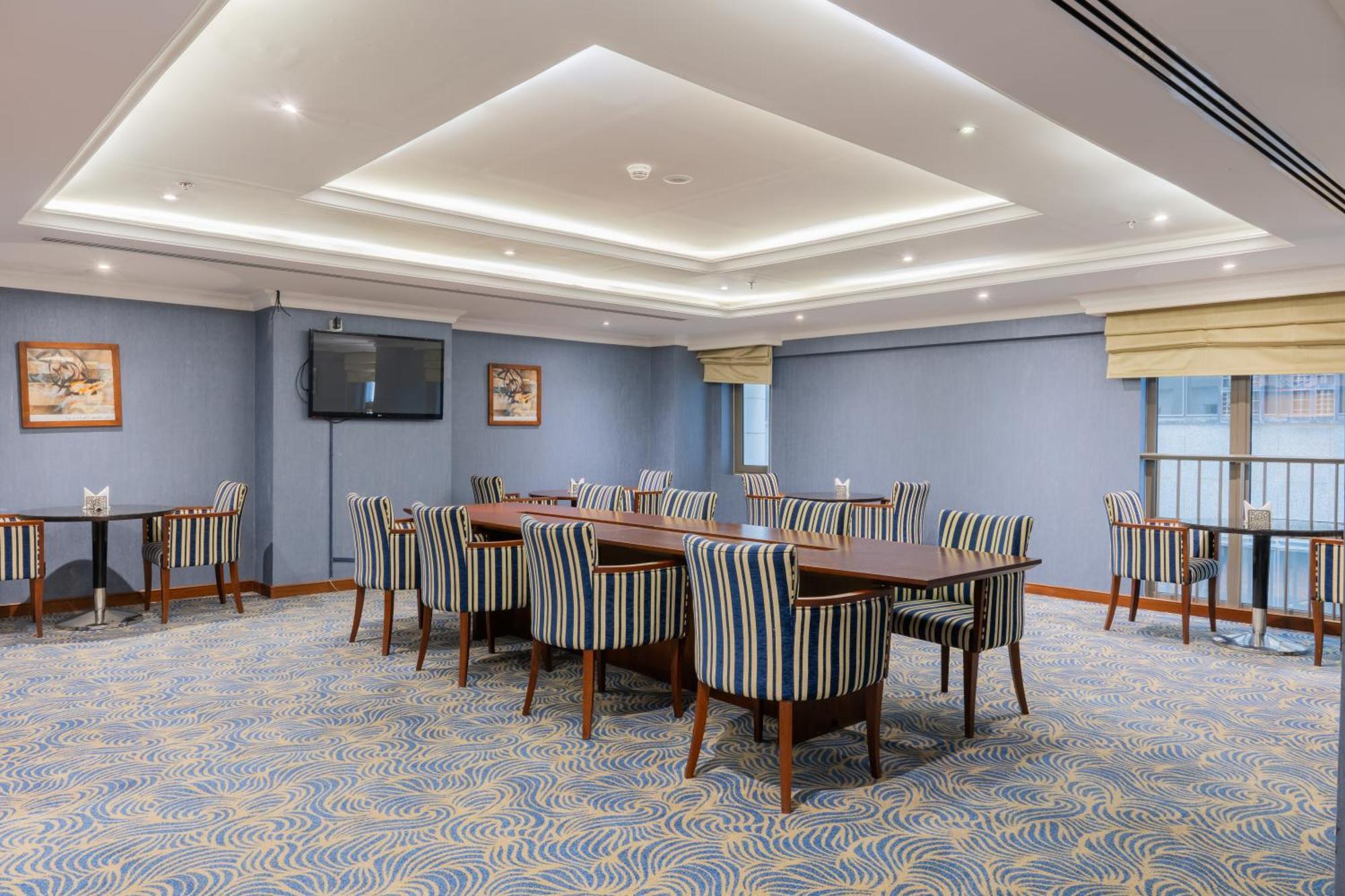 Al Aqeeq Madinah Hotel Medina Exteriör bild The photo shows a spacious conference or meeting room. It features a light blue color scheme with a patterned carpet. There are several rectangular tables arranged in a group, surrounded by striped chairs. The room is well-lit with modern ceiling lig