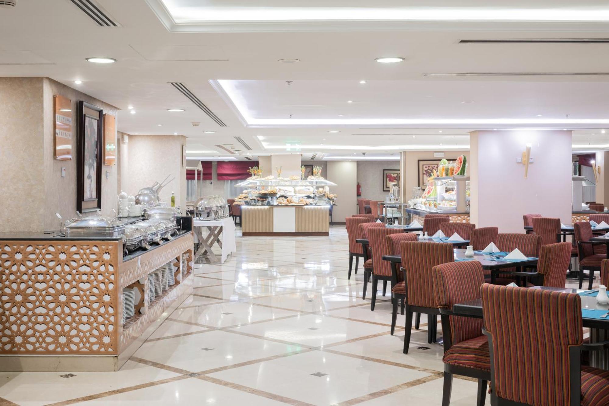 Al Aqeeq Madinah Hotel Medina Exteriör bild The photo shows an elegantly designed restaurant or dining area. It features a spacious layout with a glossy tiled floor and bright, soft lighting. There are several dining tables with striped chairs arranged neatly. Along one side, there's a buffet 