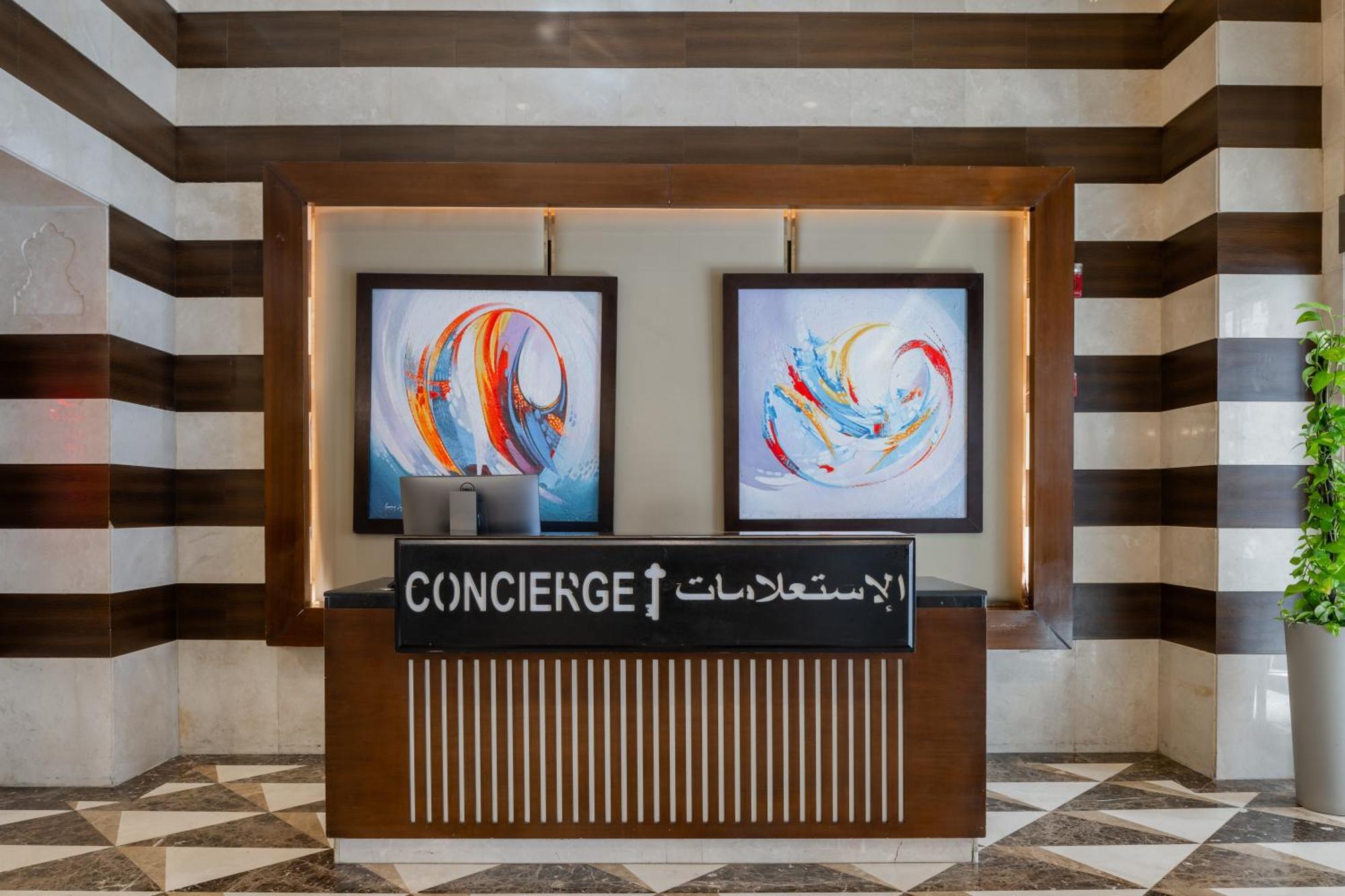 Al Aqeeq Madinah Hotel Medina Exteriör bild The image shows a concierge desk in a hotel lobby. The desk is made of wood with a striped design and features two large, colorful abstract paintings on the wall behind it. The sign on the desk reads "CONCIERGE" in English and has Arabic text below i