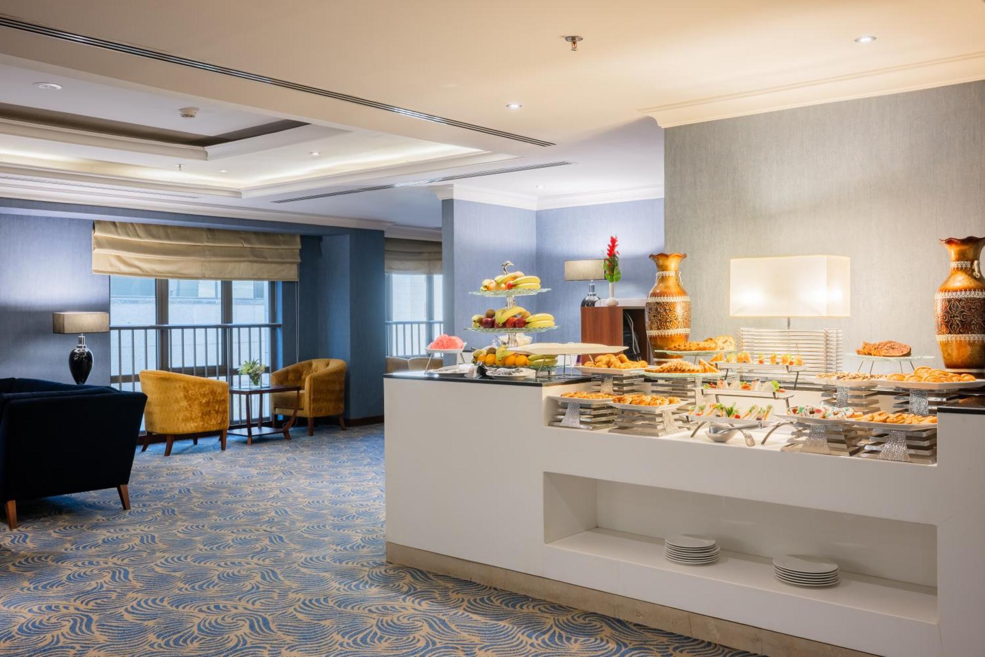 Al Aqeeq Madinah Hotel Medina Exteriör bild The photo shows a modern dining area, likely in a hotel or restaurant. On the right side, there is a buffet table with various food items displayed, including pastries and desserts. Behind the table, a decorative fountain is visible. The room feature