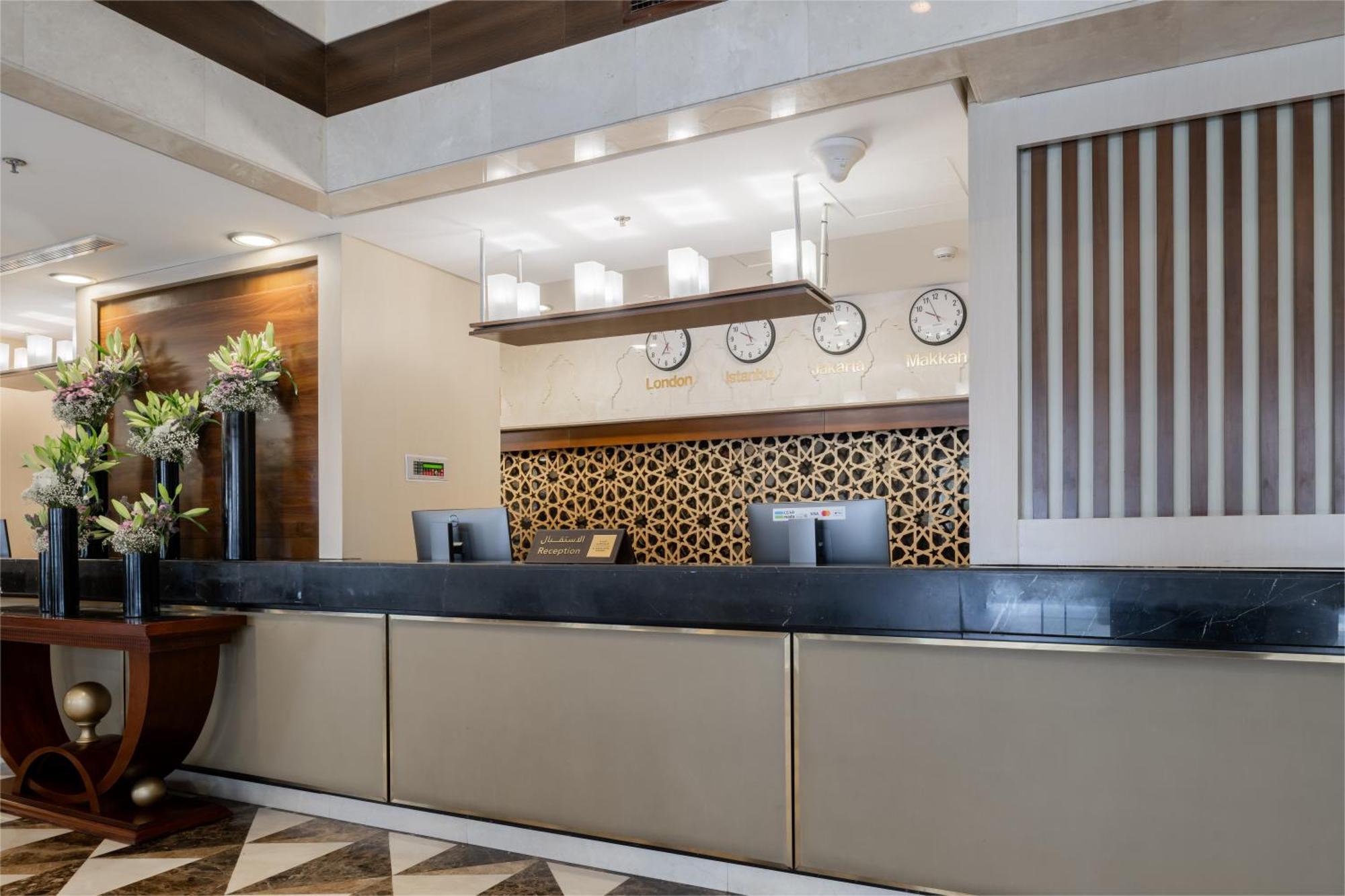 Al Aqeeq Madinah Hotel Medina Exteriör bild The photo shows the interior of a hotel reception area. The reception desk is designed with a sleek, modern look featuring a dark countertop and a light-colored front. Above the desk, there are several clocks displaying different times, suggesting an