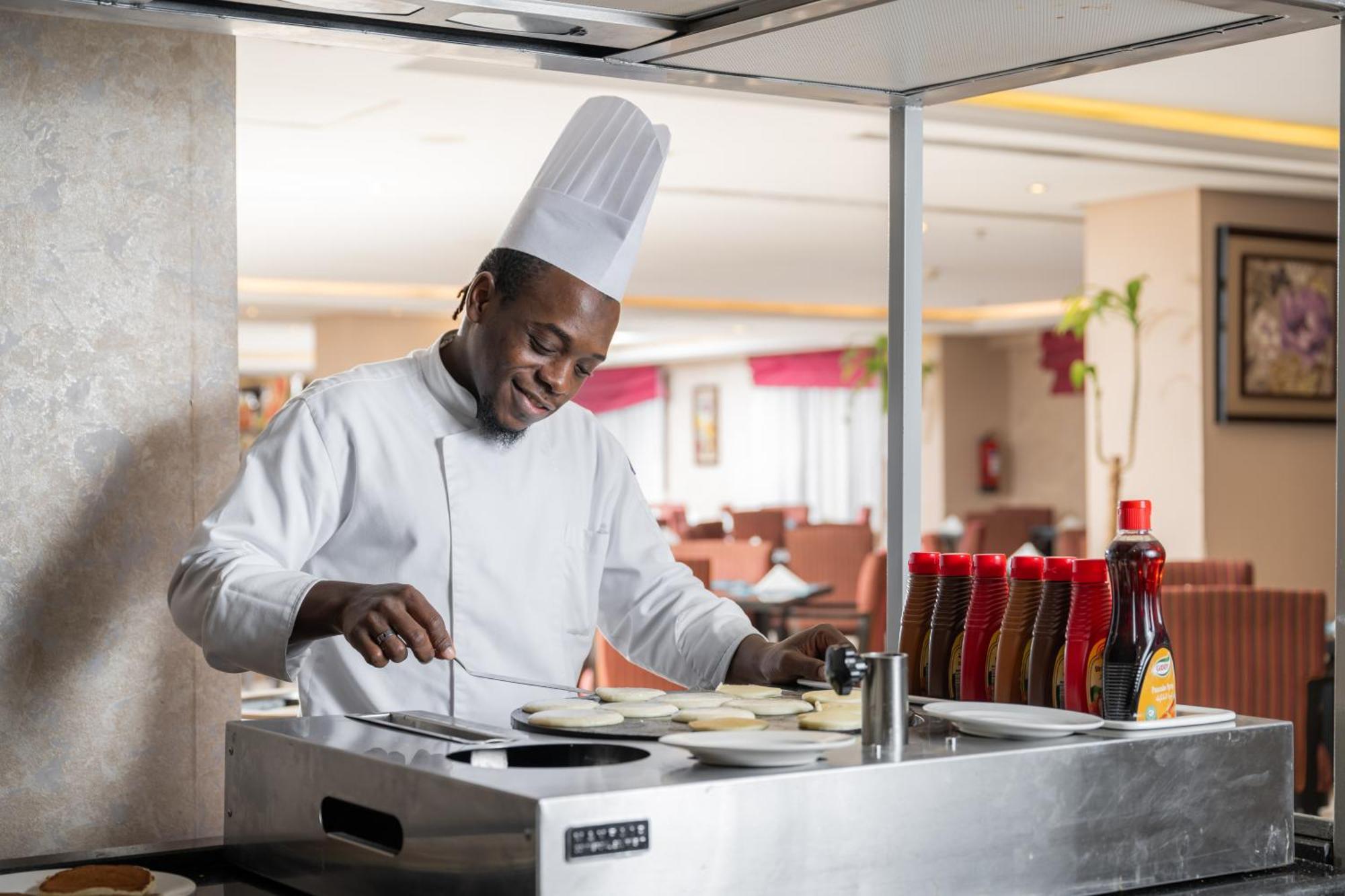 Al Aqeeq Madinah Hotel Medina Exteriör bild The photo shows a chef in a professional kitchen setting. He is wearing a traditional white chef's uniform and a tall white chef's hat, and he appears to be smiling while cooking. The chef is standing at a stainless steel cooking station, where he is