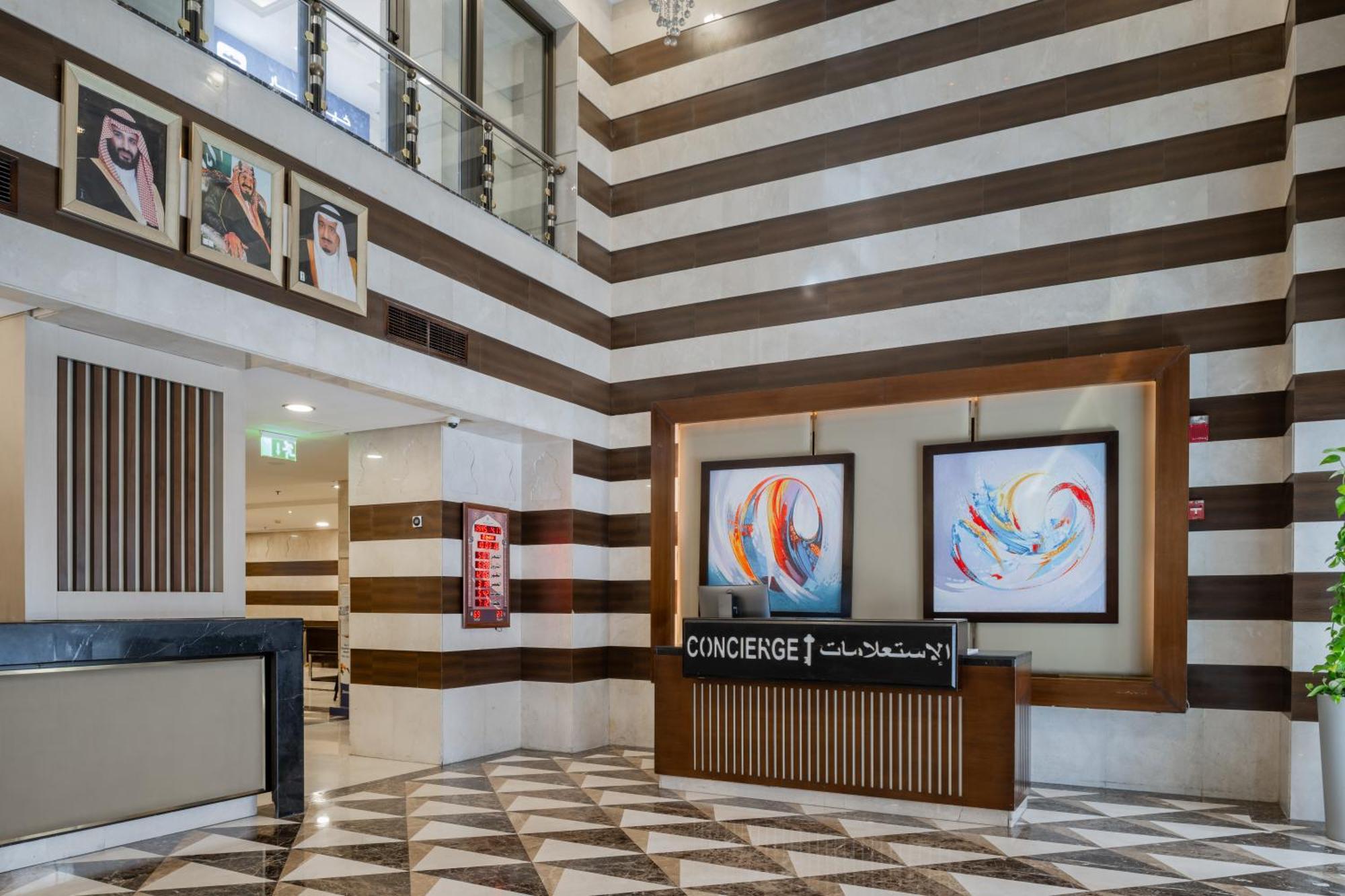 Al Aqeeq Madinah Hotel Medina Exteriör bild The photo shows a modern hotel lobby area characterized by its stylish design. The walls feature a pattern of horizontal stripes in different shades, creating a sophisticated atmosphere. To the left, there is a reception area with a sleek desk labele