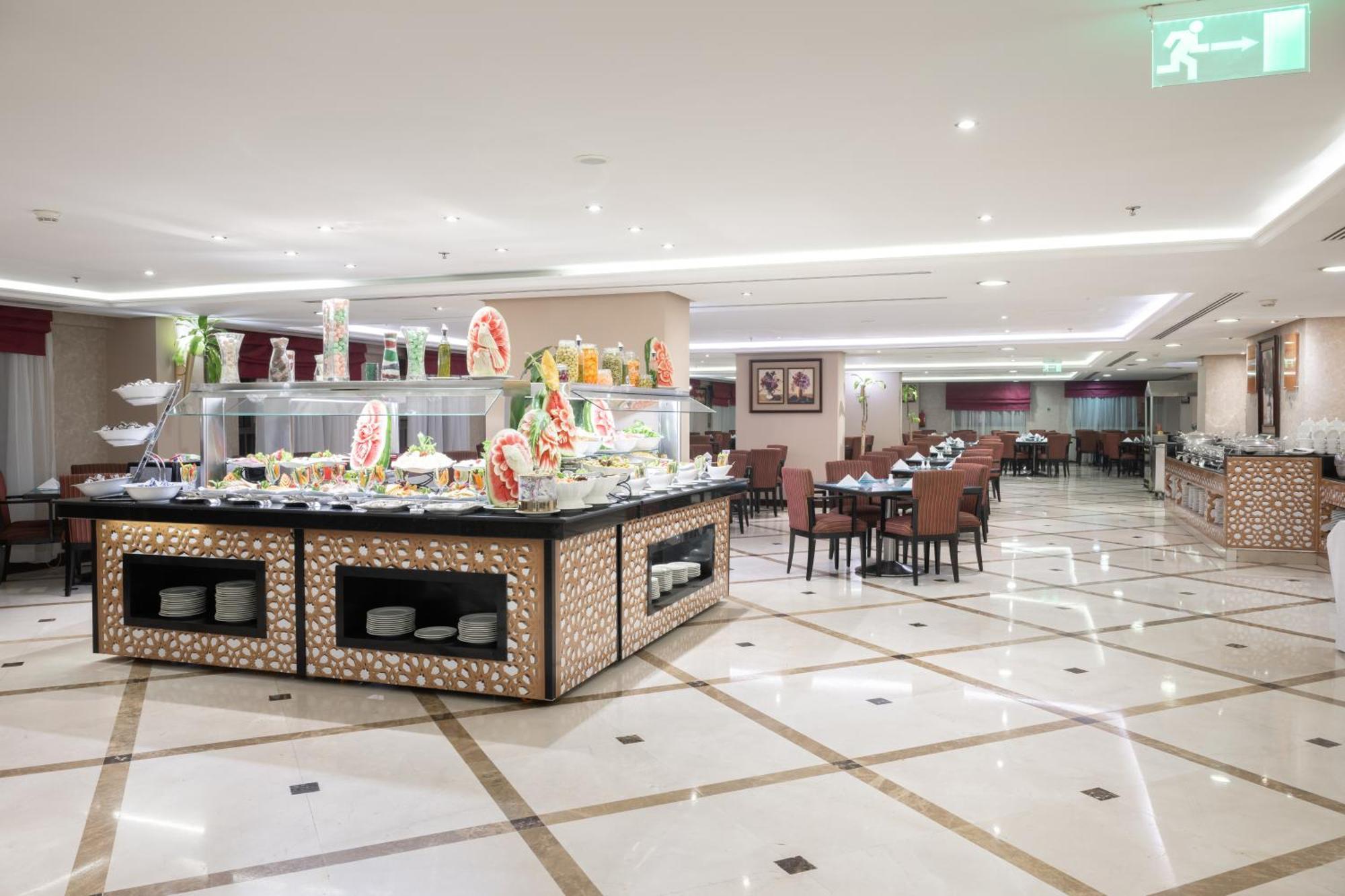 Al Aqeeq Madinah Hotel Medina Exteriör bild The photo shows a spacious restaurant interior with a modern design. In the foreground, there is a large buffet counter filled with various food items, beautifully arranged and decorated. The counter features a backlit display, showcasing fruits and 