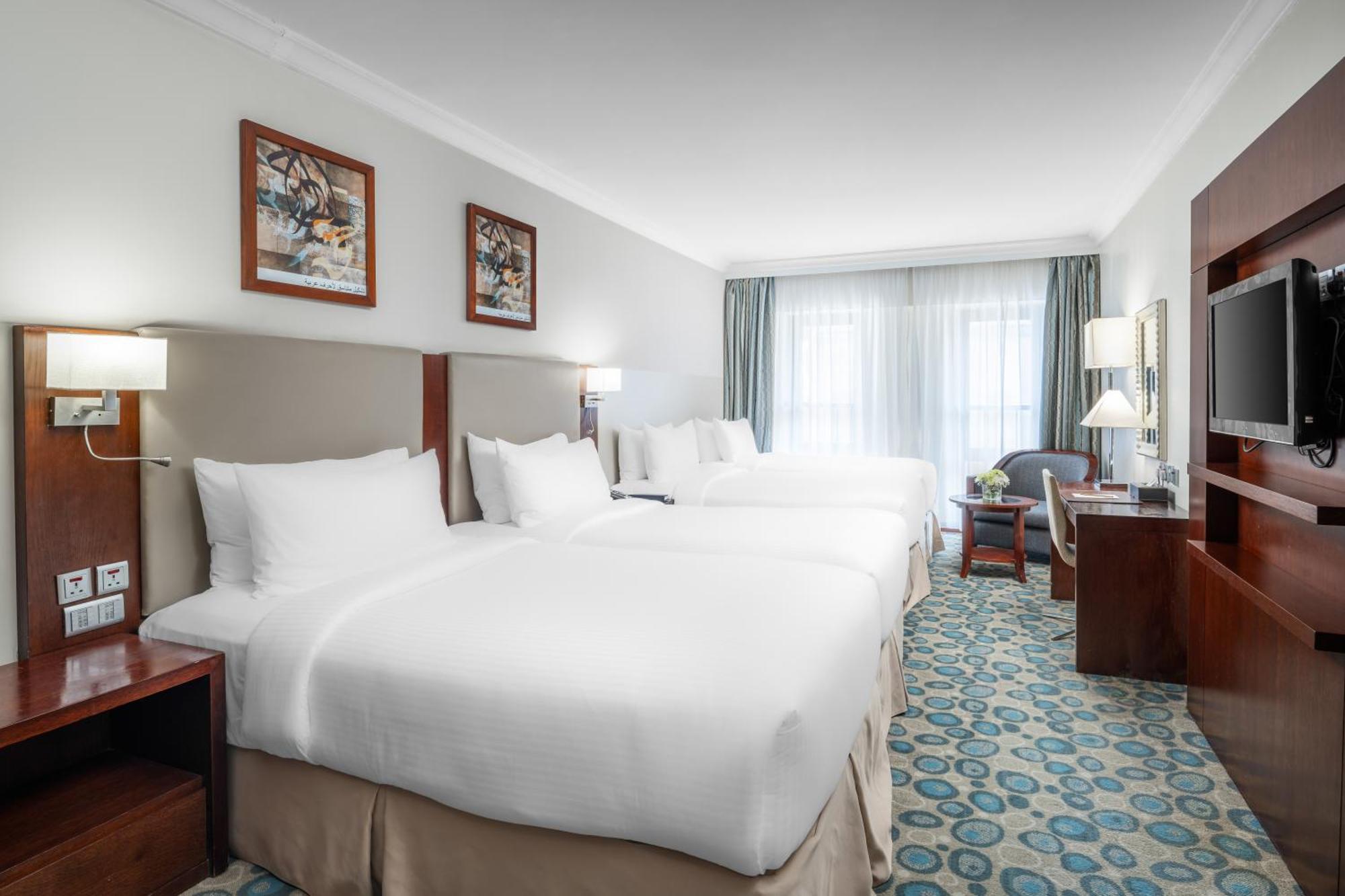 Al Aqeeq Madinah Hotel Medina Exteriör bild The photo shows a hotel room featuring a modern, clean design. There are two large beds with white linens positioned side by side. Above the beds are framed artworks, and soft lighting is provided by wall sconces. To the right, there is a wooden ente