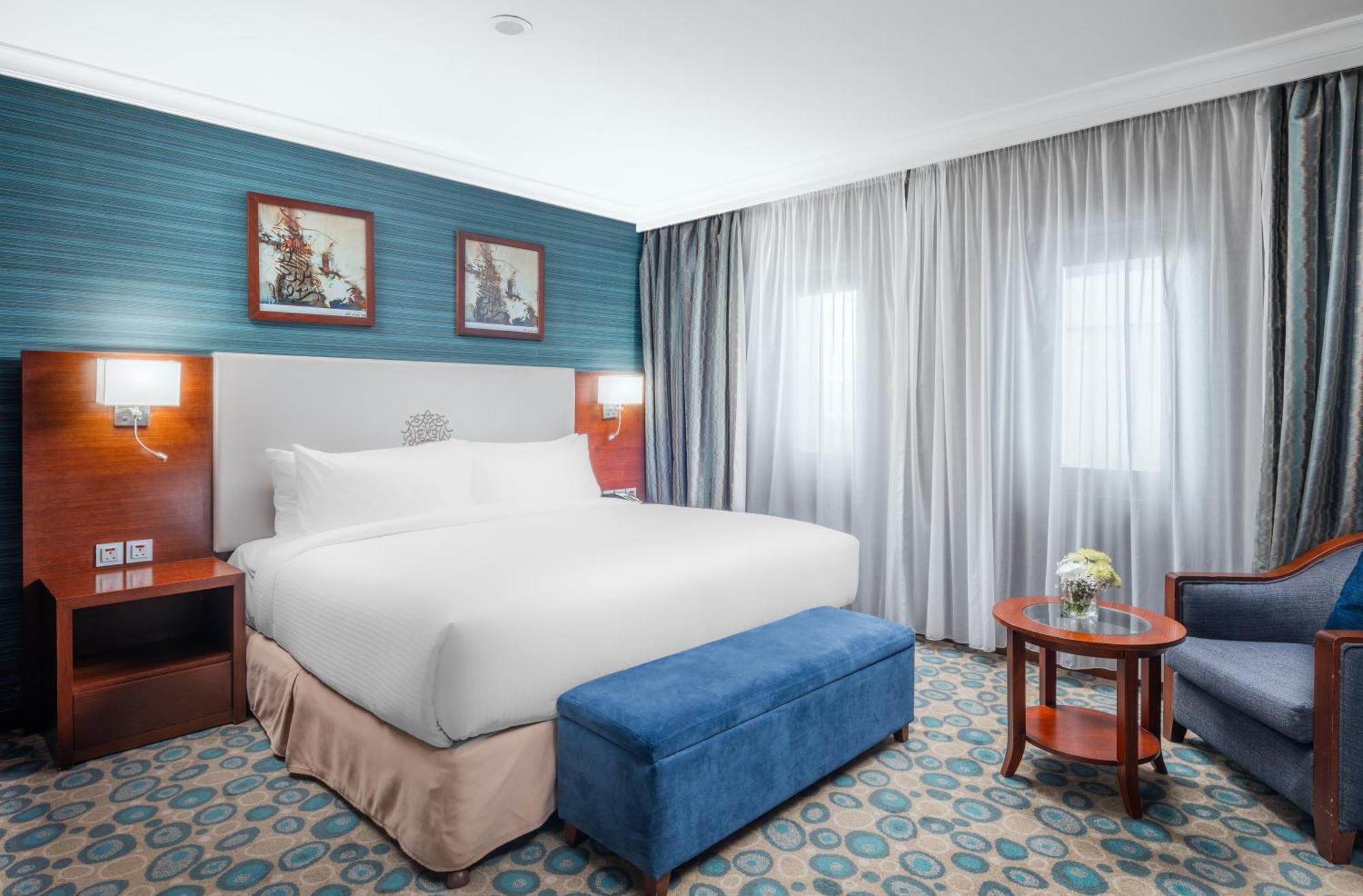 Al Aqeeq Madinah Hotel Medina Exteriör bild The photo shows a hotel room featuring a large, neatly made bed with a white duvet. The walls have a light blue accent with framed artwork above the bed. There’s a blue ottoman at the foot of the bed. To one side, there’s a small wooden table with a 