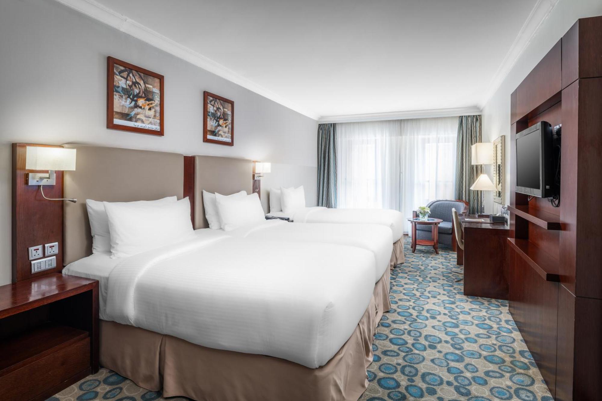 Al Aqeeq Madinah Hotel Medina Exteriör bild The photo shows a well-furnished hotel room featuring two large beds with white bedding. The walls are adorned with framed artwork. There are bedside lamps next to each bed, and a flat-screen TV is mounted on the wall. A large window allows natural l
