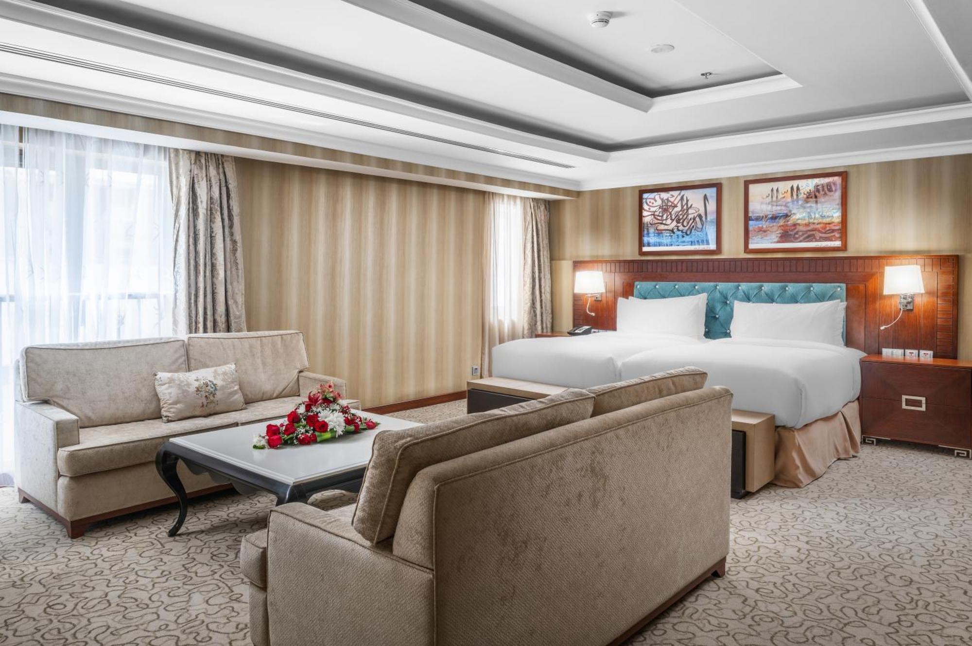 Al Aqeeq Madinah Hotel Medina Exteriör bild The photo shows a well-decorated hotel room. It features a large bed with two pillows and a light-colored duvet. There are two comfortable couches arranged around a coffee table with a floral centerpiece. The walls have a subtle striped design, and l