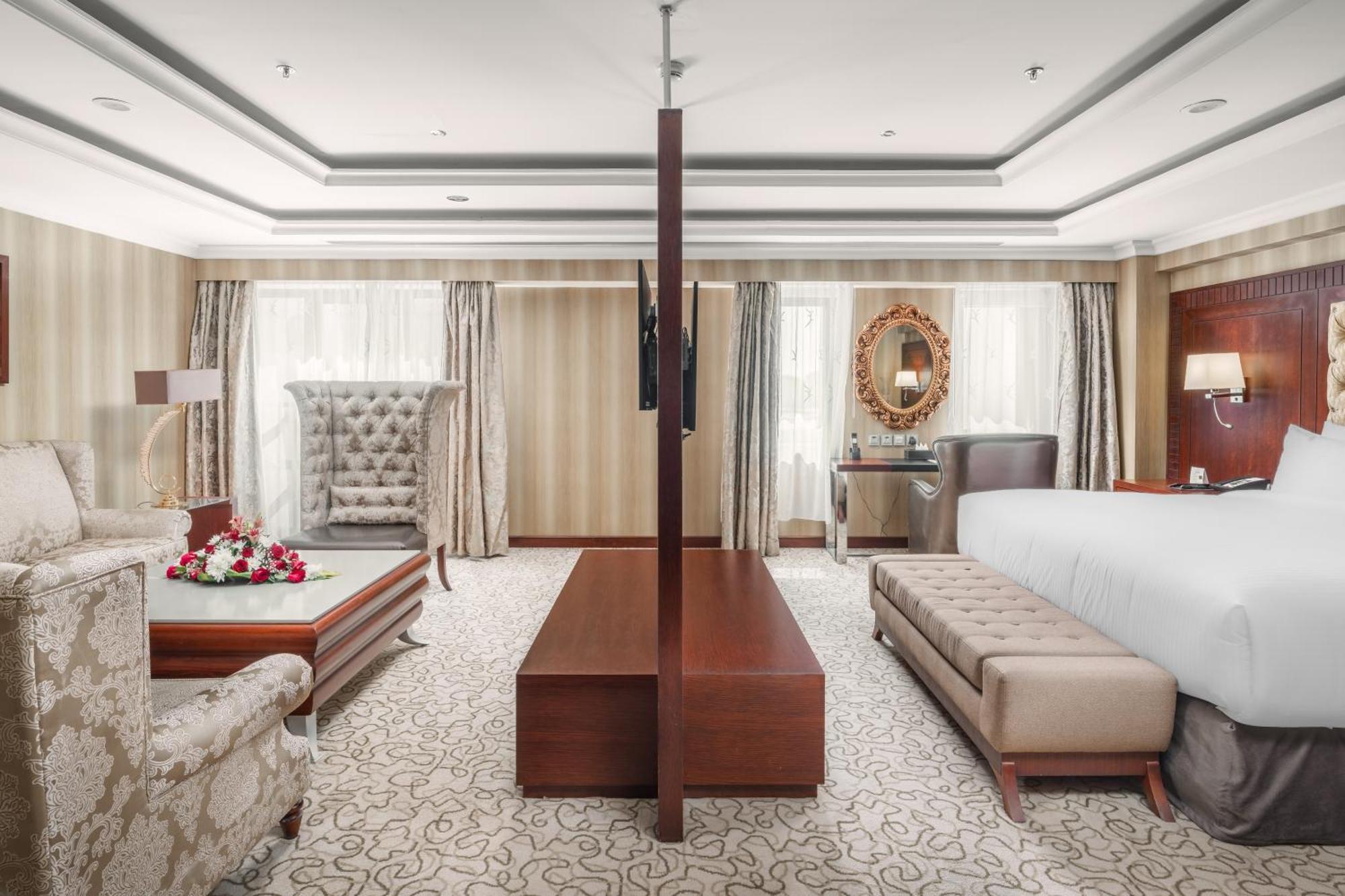 Al Aqeeq Madinah Hotel Medina Exteriör bild The photo showcases a spacious hotel suite with an elegant and modern design. The room features a large bed on one side, complemented by a plush bench at the foot. Opposite the bed, there is a stylish seating area that includes a decorative armchair 