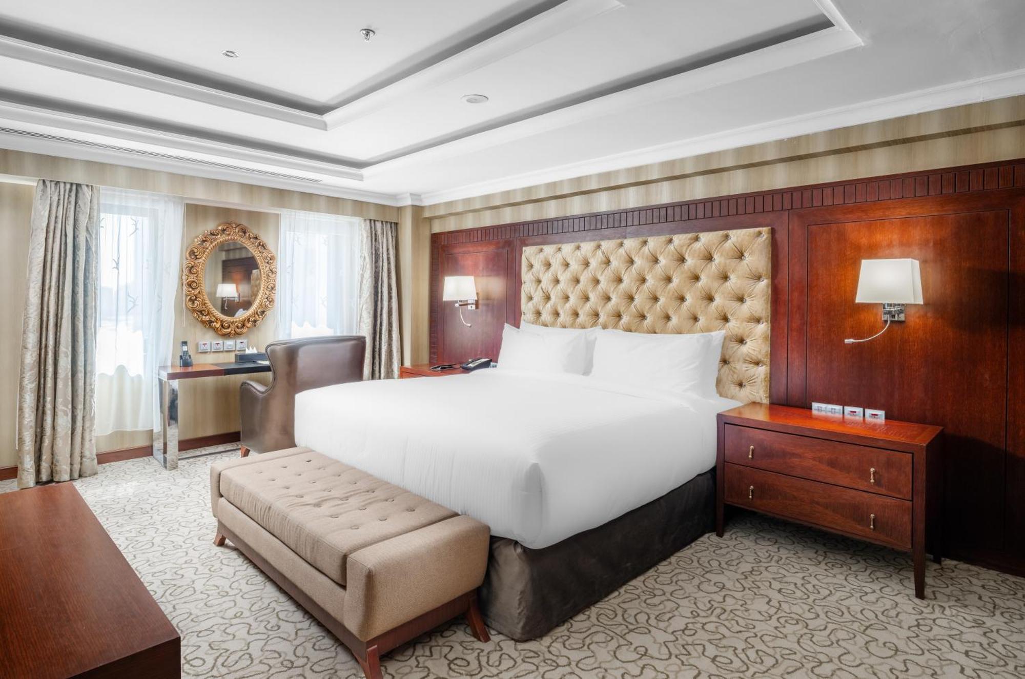Al Aqeeq Madinah Hotel Medina Exteriör bild The photo shows a well-furnished hotel room featuring a large bed with a tufted headboard. The room has a warm color palette with wooden accents and soft lighting. There are two windows with curtains that allow natural light to fill the space. Alongs