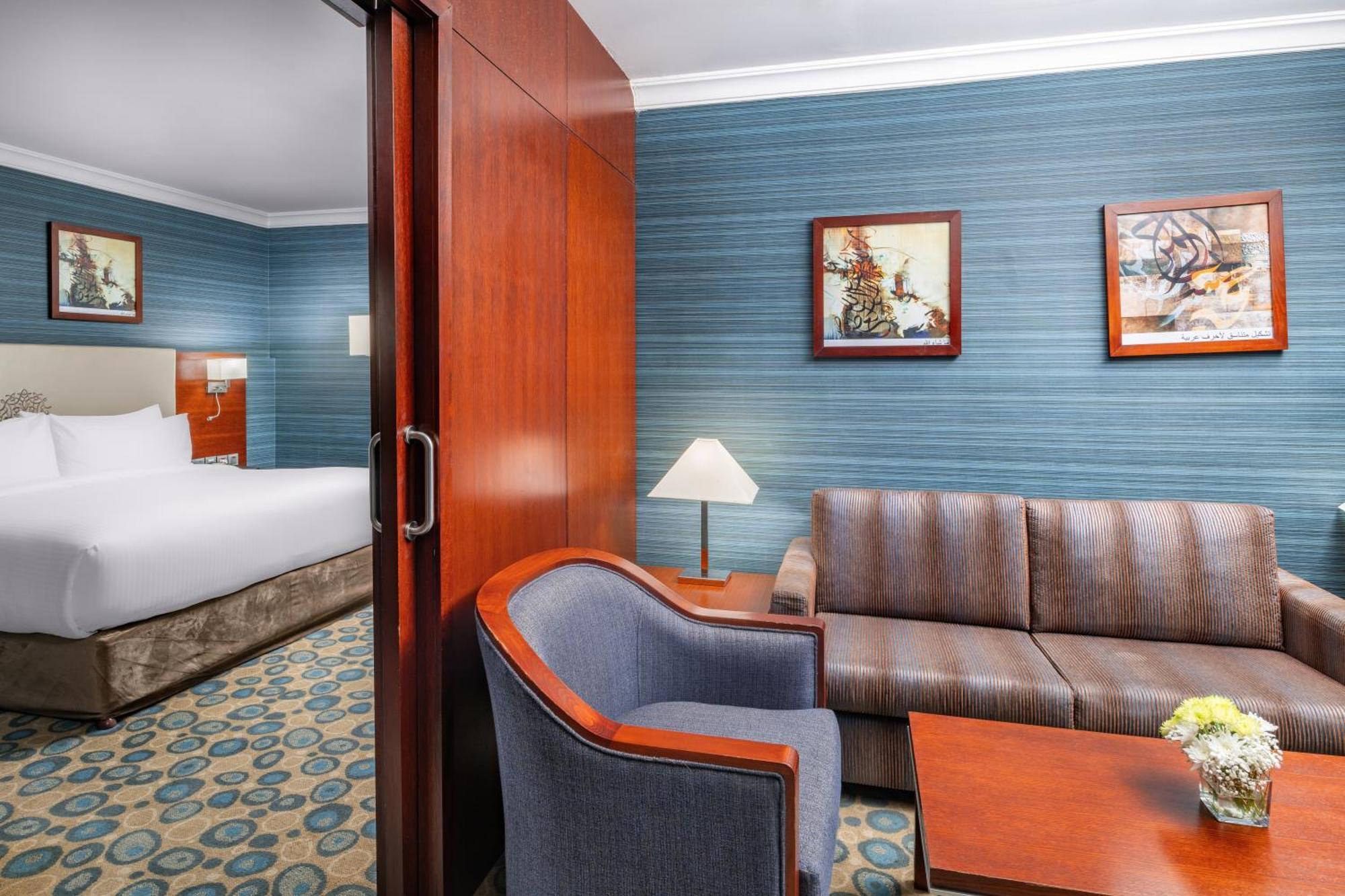 Al Aqeeq Madinah Hotel Medina Exteriör bild The photo shows a well-furnished hotel room featuring a living area and a bedroom. To the left, there is a bed visible through an open doorway. The living area includes a gray upholstered chair and a brown couch, both arranged around a small table. A