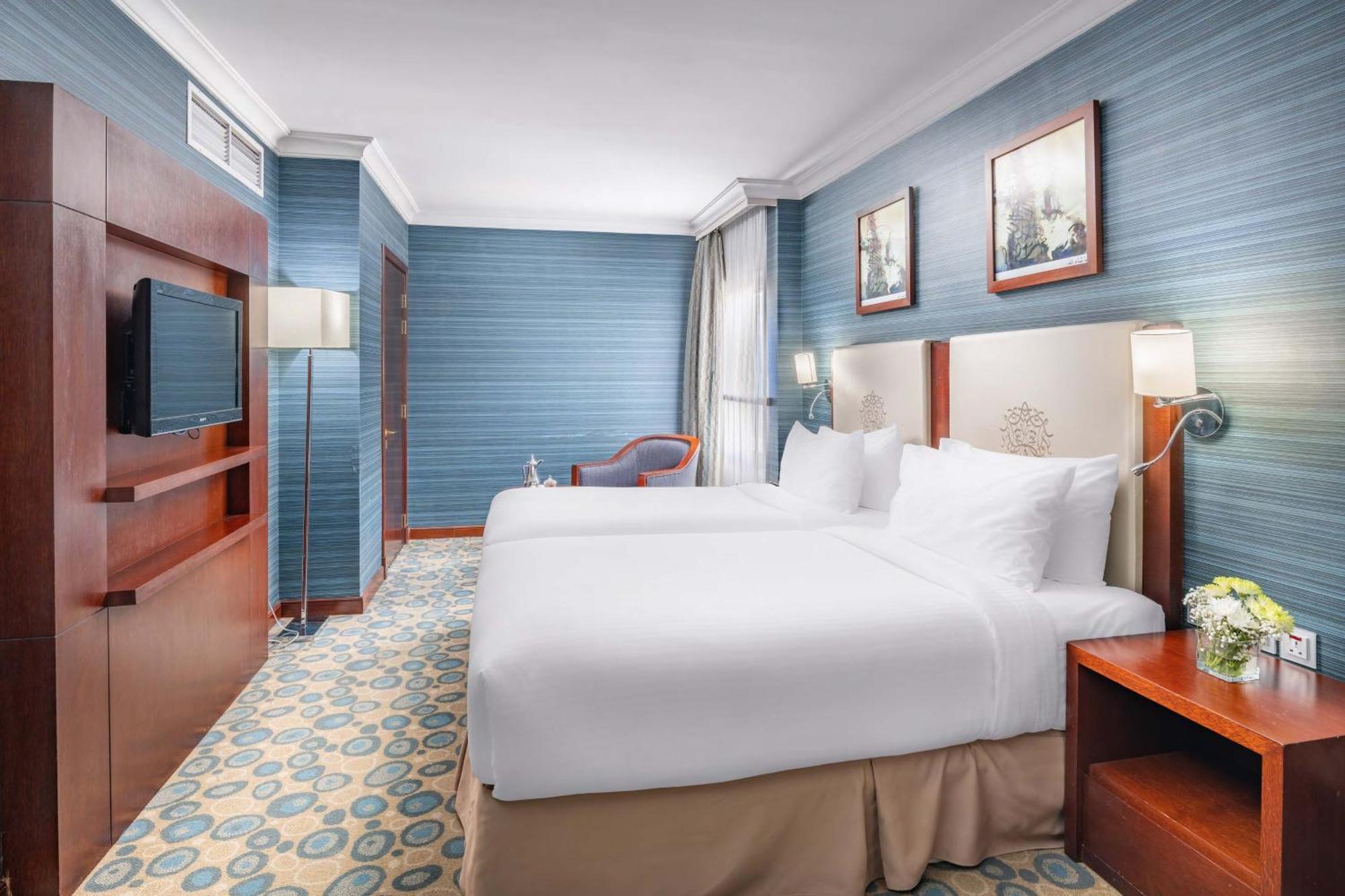 Al Aqeeq Madinah Hotel Medina Exteriör bild The photo shows a hotel room featuring two twin beds dressed in white linens. The walls are painted in a soft blue color, and there are framed artworks hung above the beds. A flat-screen TV is mounted on a wooden unit against one wall. To the side, t