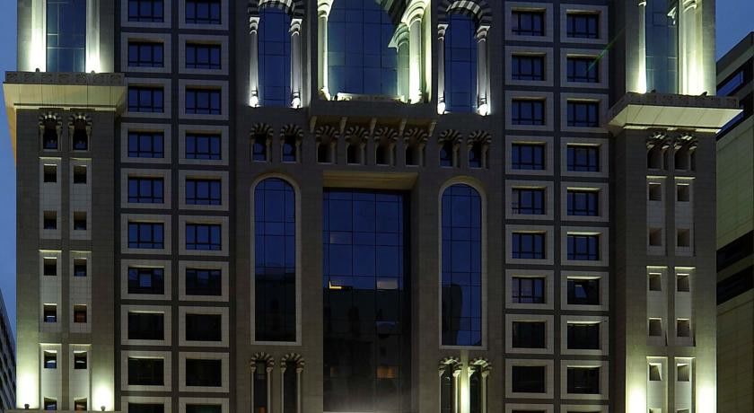 Al Aqeeq Madinah Hotel Medina Exteriör bild The image shows the facade of a modern high-rise building illuminated at night. The design features large glass windows arranged in a grid pattern, along with decorative architectural elements at the top, including arches and ornate detailing. The co