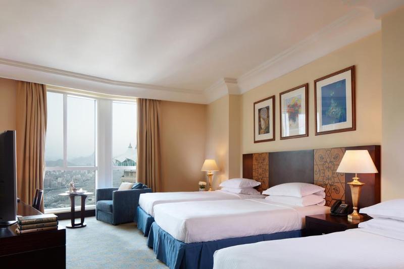 Al Aqeeq Madinah Hotel Medina Exteriör bild The photo shows a hotel room featuring three beds arranged neatly. The room has a warm, inviting ambiance with beige walls and a soft carpet. Large windows allow natural light to fill the space, offering a view outside. There are curtains in a light 