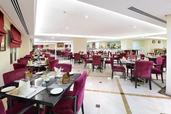 Al Aqeeq Madinah Hotel Medina Exteriör bild The photo shows an elegant restaurant interior with a spacious layout. The dining area features multiple tables, each set with white tablecloths and dinnerware. The chairs are upholstered in a rich purple fabric, adding a touch of luxury to the atmos