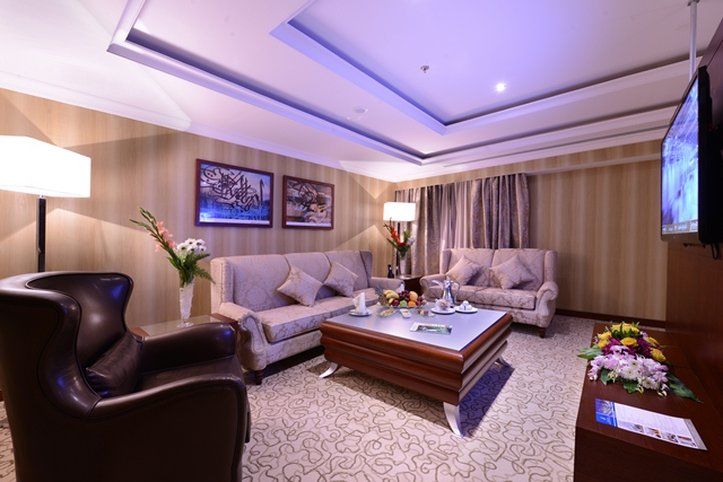 Al Aqeeq Madinah Hotel Medina Exteriör bild The photo shows a stylish and comfortable living room. It features two light-colored sofas with decorative cushions arranged around a modern coffee table. In one corner, there's a dark, leather armchair. The walls are adorned with framed artwork, and