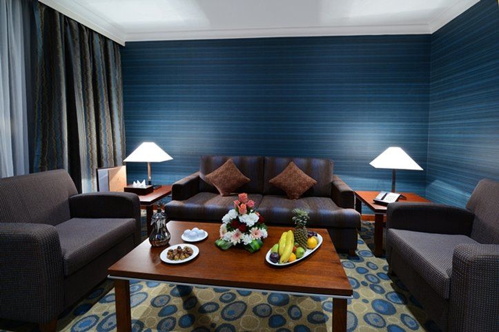 Al Aqeeq Madinah Hotel Medina Exteriör bild The photo shows a cozy living area featuring a dark blue wall with a textured design. In the center, there is a wooden coffee table surrounded by two comfortable armchairs and a sofa with decorative cushions. On the table, there are plates with snack