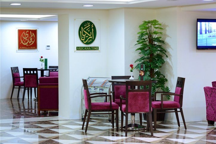 Al Aqeeq Madinah Hotel Medina Exteriör bild The photo shows an interior space, likely a dining or lounge area. There are several tables with four chairs each, featuring a modern design with dark wood and plush maroon upholstery. On the walls, there are decorative signs, one of which has Arabic