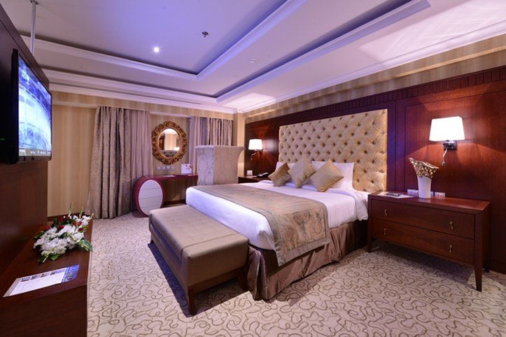 Al Aqeeq Madinah Hotel Medina Exteriör bild The photo shows an elegantly designed hotel room. It features a large bed with a plush headboard and decorative pillows. On either side of the bed are bedside tables with lamps. There's a comfortable bench at the foot of the bed and a TV mounted on t
