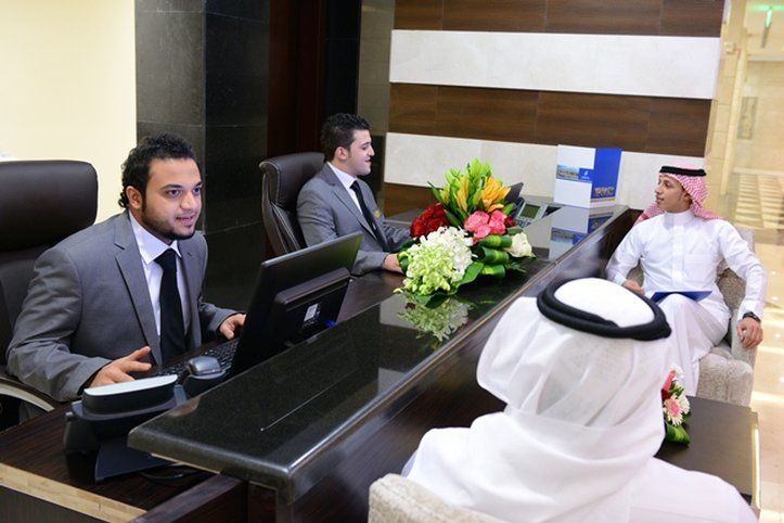 Al Aqeeq Madinah Hotel Medina Exteriör bild The photo depicts a reception area with three individuals. Two men are seated behind a reception desk; they are dressed in formal suits and appear to be engaged in conversation, possibly assisting a guest. The desk has a computer and a vibrant arrang