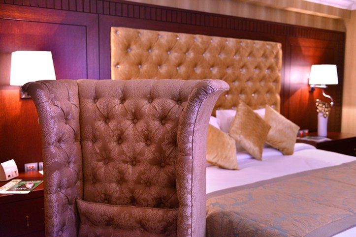Al Aqeeq Madinah Hotel Medina Exteriör bild The image shows a luxurious bedroom featuring a large upholstered headboard with a tufted design in a soft, beige color. The bed is elegantly dressed with decorative pillows and a textured bedspread. In the background, there are wooden paneling and s